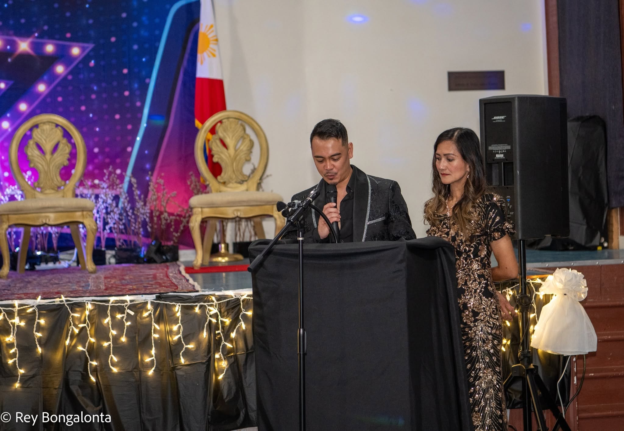 Ajay Delarosa speaking at a formal Filipino community gala with the Philippine flag in the background